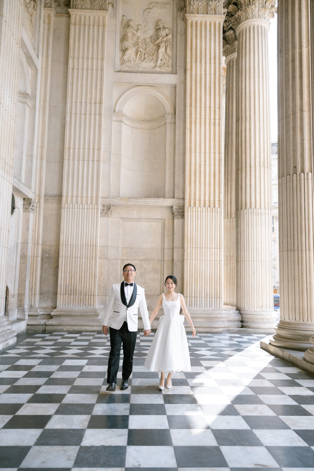 A couple in formal wedding attire walks hand in hand across a grand, checkered marble floor with tall columns and ornate walls in the background.