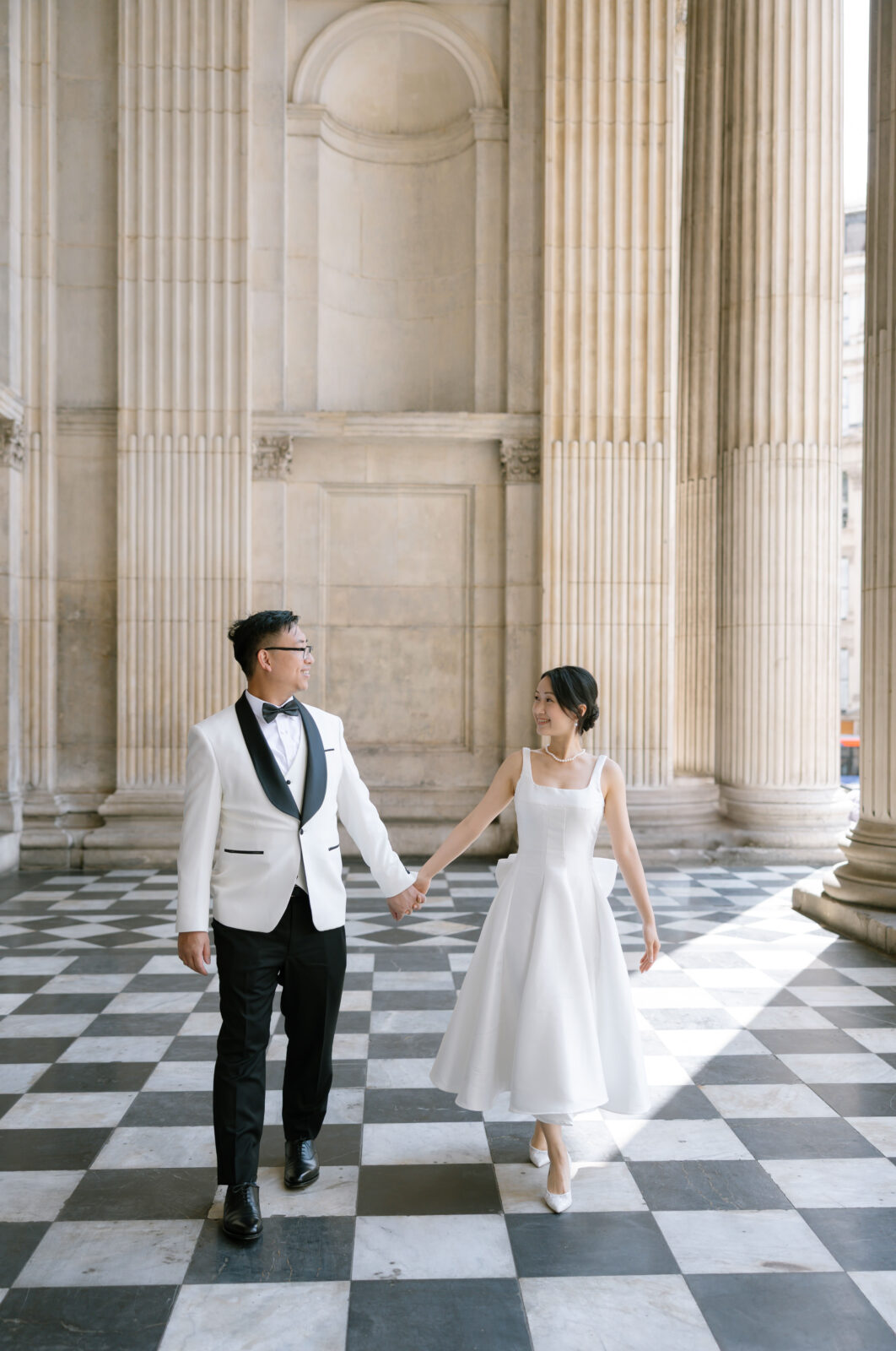 A couple dressed in formal wedding attire, holding hands and smiling at each other, walk on a black-and-white checkered floor between tall stone columns in a grand, classical building.