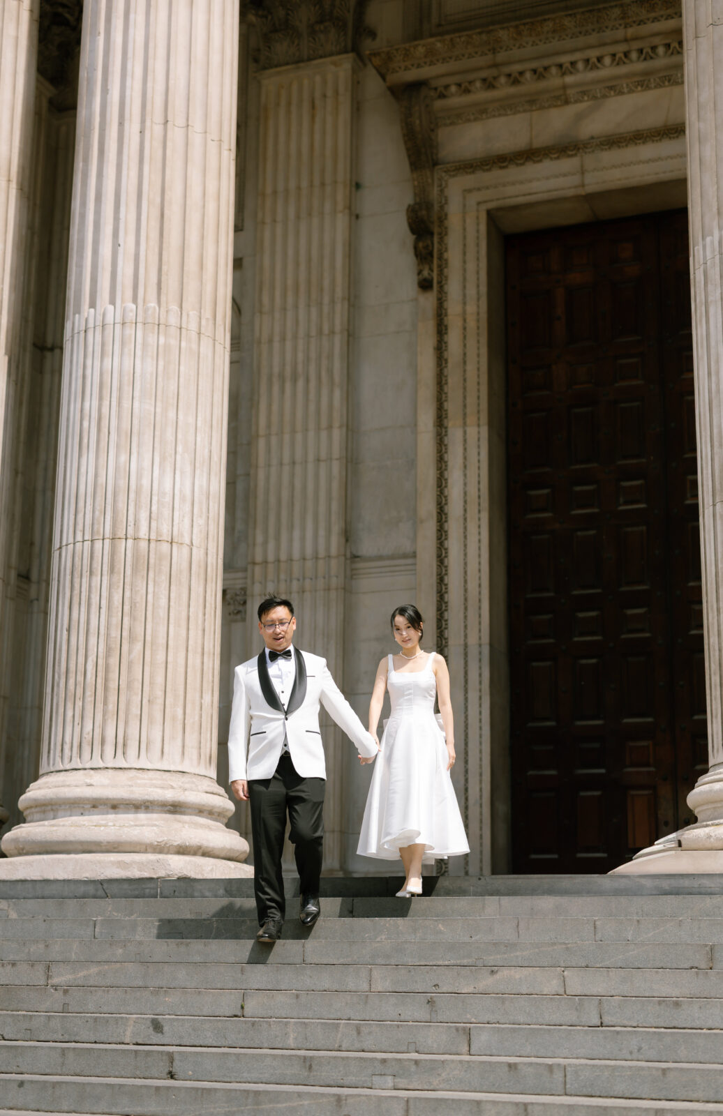 A couple in formal attire, with the man in a white suit and the woman in a white dress, walk down stone steps outside a grand building with tall columns and a large wooden door.