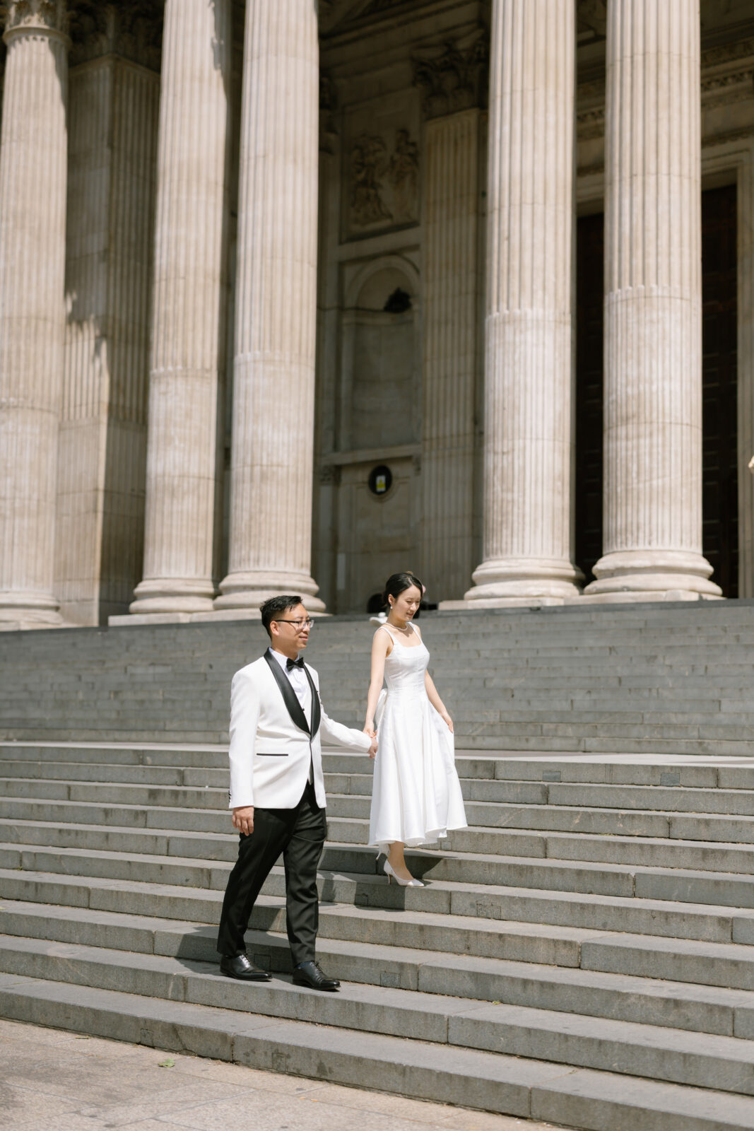 A couple dressed in formal attire walks hand in hand down stone steps in front of a grand building with tall columns. The man wears a white tuxedo jacket and the woman wears a white dress.