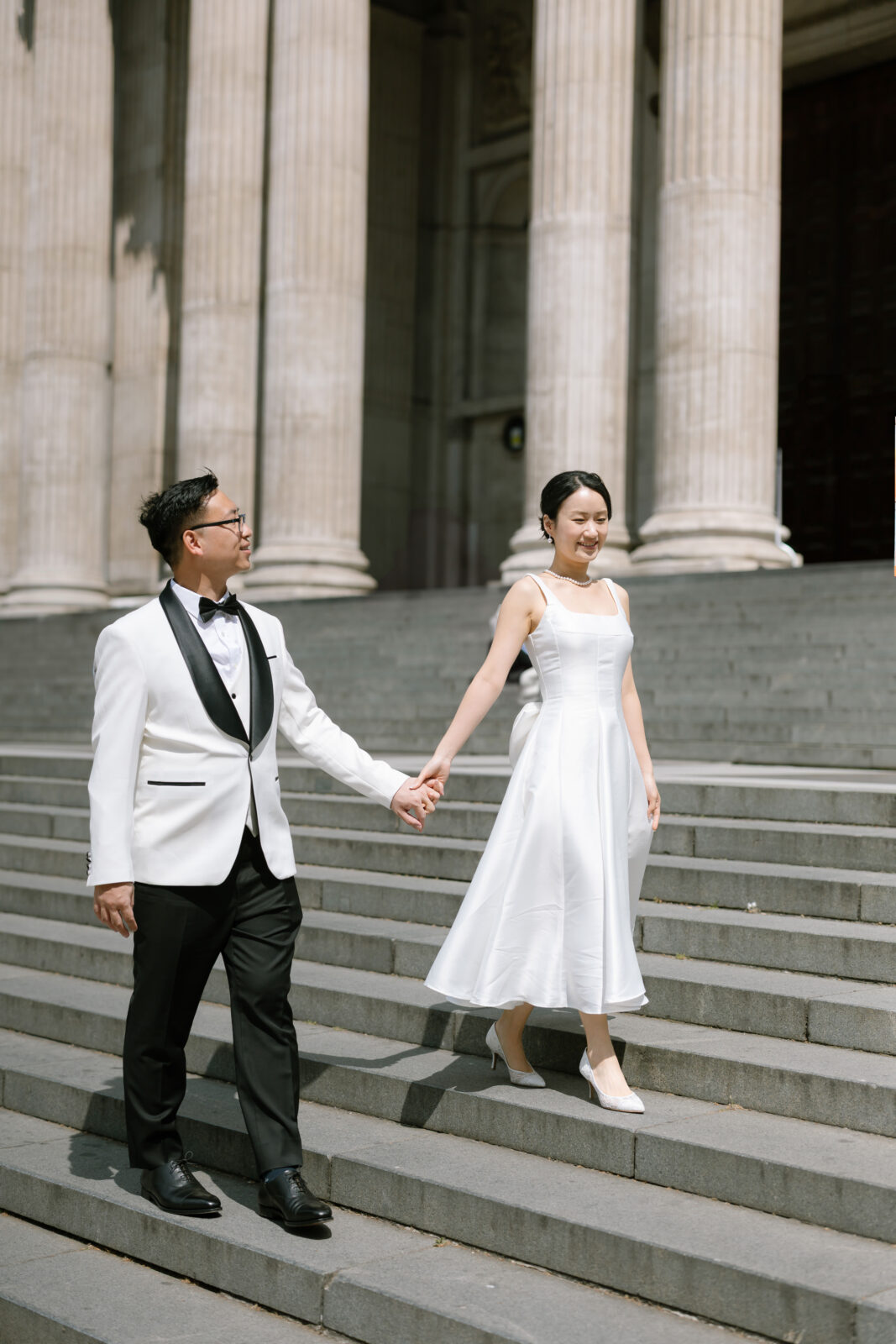 A couple dressed elegantly in black and white formal attire holds hands while walking down stone steps in front of a grand building with tall columns.