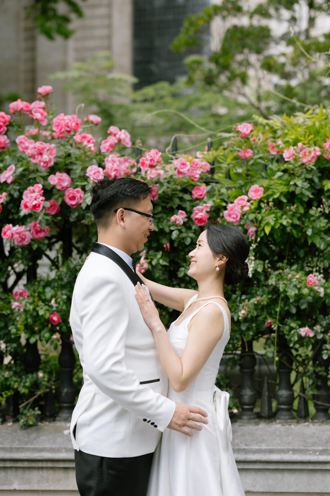 A couple dressed in formal wedding attire smiles at each other in front of lush green bushes with vibrant pink roses outdoors. The groom wears a white suit and the bride wears a white dress and pearls.