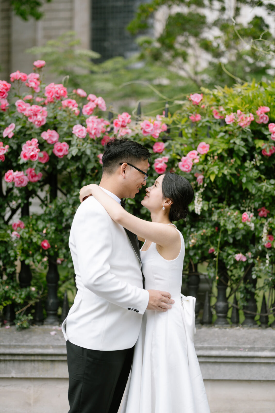 A couple dressed in white wedding attire embrace and smile at each other in front of a lush green bush with vibrant pink flowers.