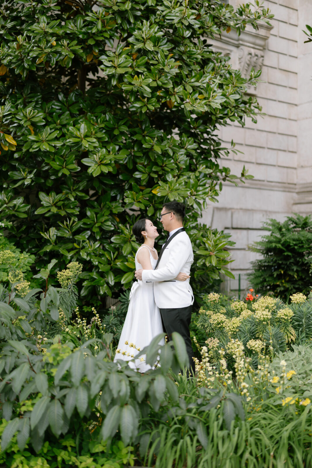 A bride and groom stand in lush greenery, gazing lovingly at each other and embracing, with a large leafy tree and a stone building in the background.