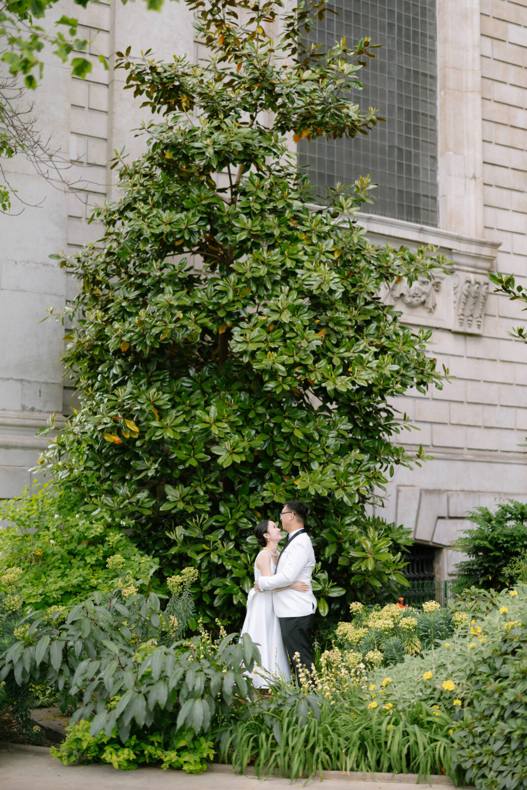 A couple in wedding attire embraces in a lush green garden, surrounded by large leafy plants and standing in front of a tall tree and a stone building.