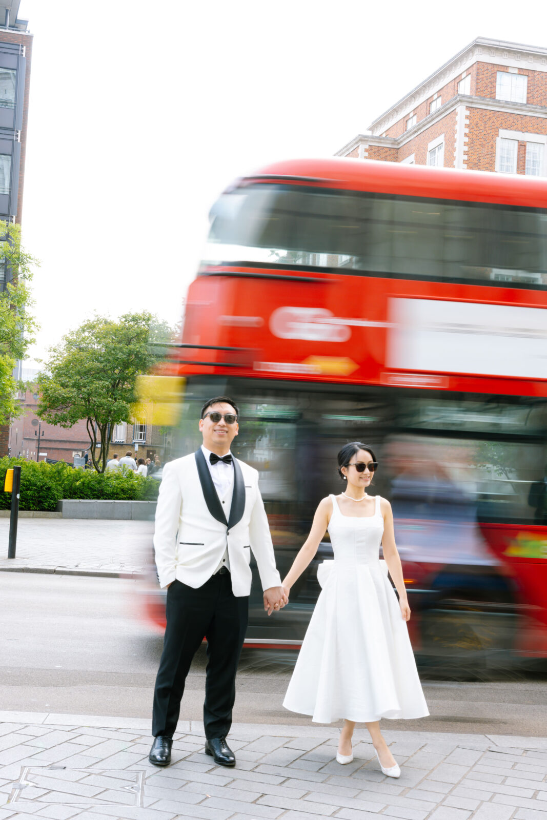 A couple dressed in formal white attire holds hands on a city street, with a red double-decker bus speeding by behind them, creating a blur effect. Trees and brick buildings are visible in the background.