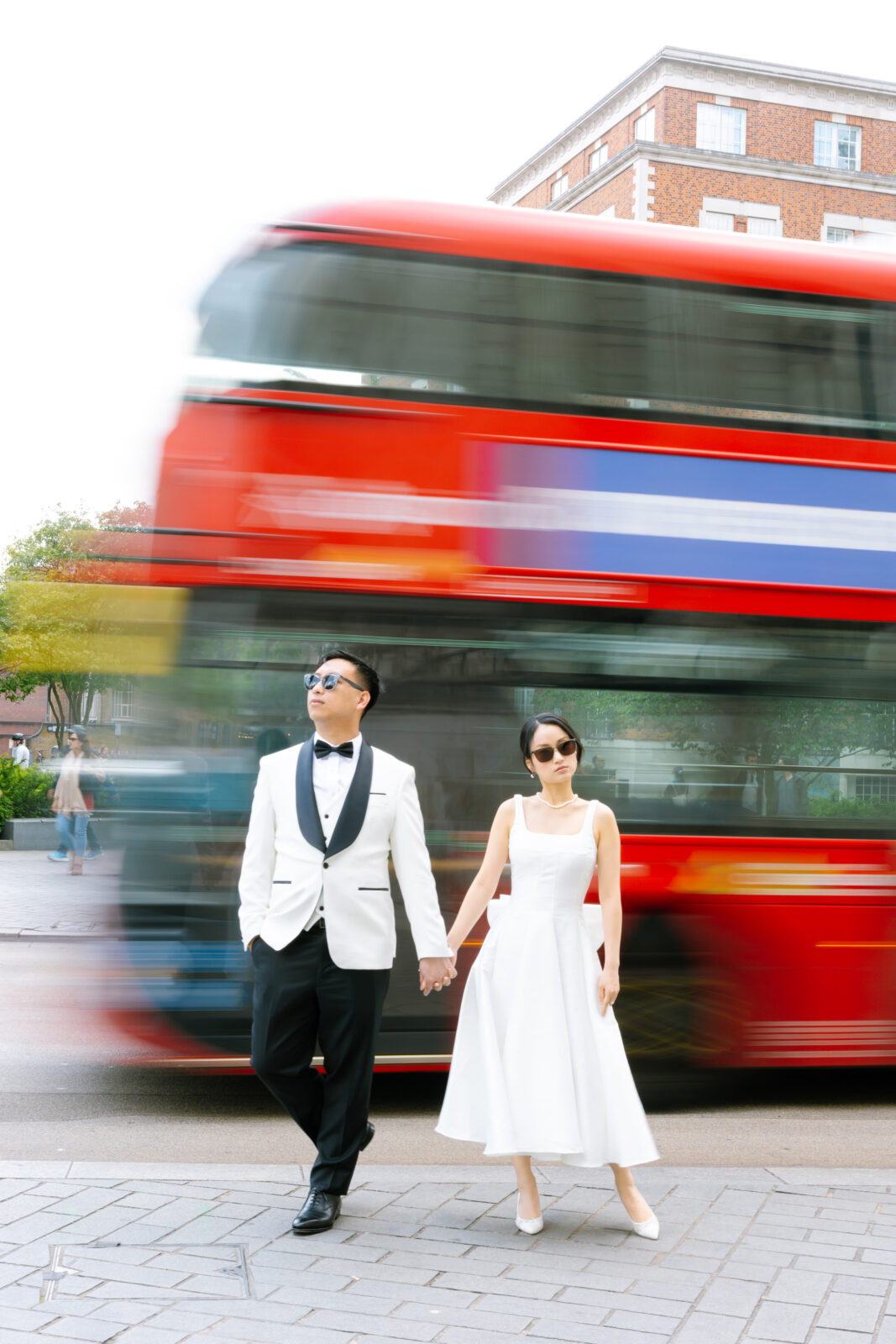 A couple in formal attire, holding hands and wearing sunglasses, stand on a city sidewalk as a red double-decker bus speeds by, creating a blurred effect in the background.