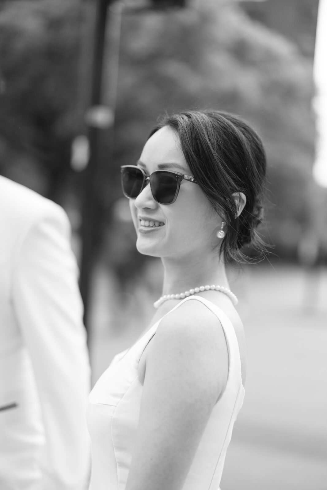 A woman in a white dress and sunglasses smiles outdoors. She wears a pearl necklace and earrings, and her hair is styled up. The image is in black and white.