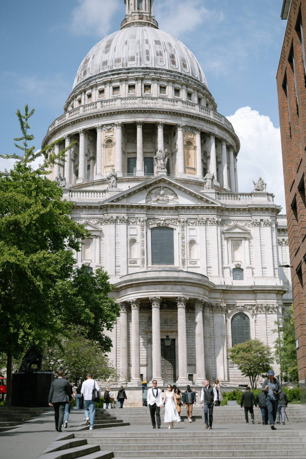 People walk near the grand entrance of St. Paul’s Cathedral in London on a sunny day, with the large dome and classical columns prominent in the background. Trees and modern buildings flank the scene.