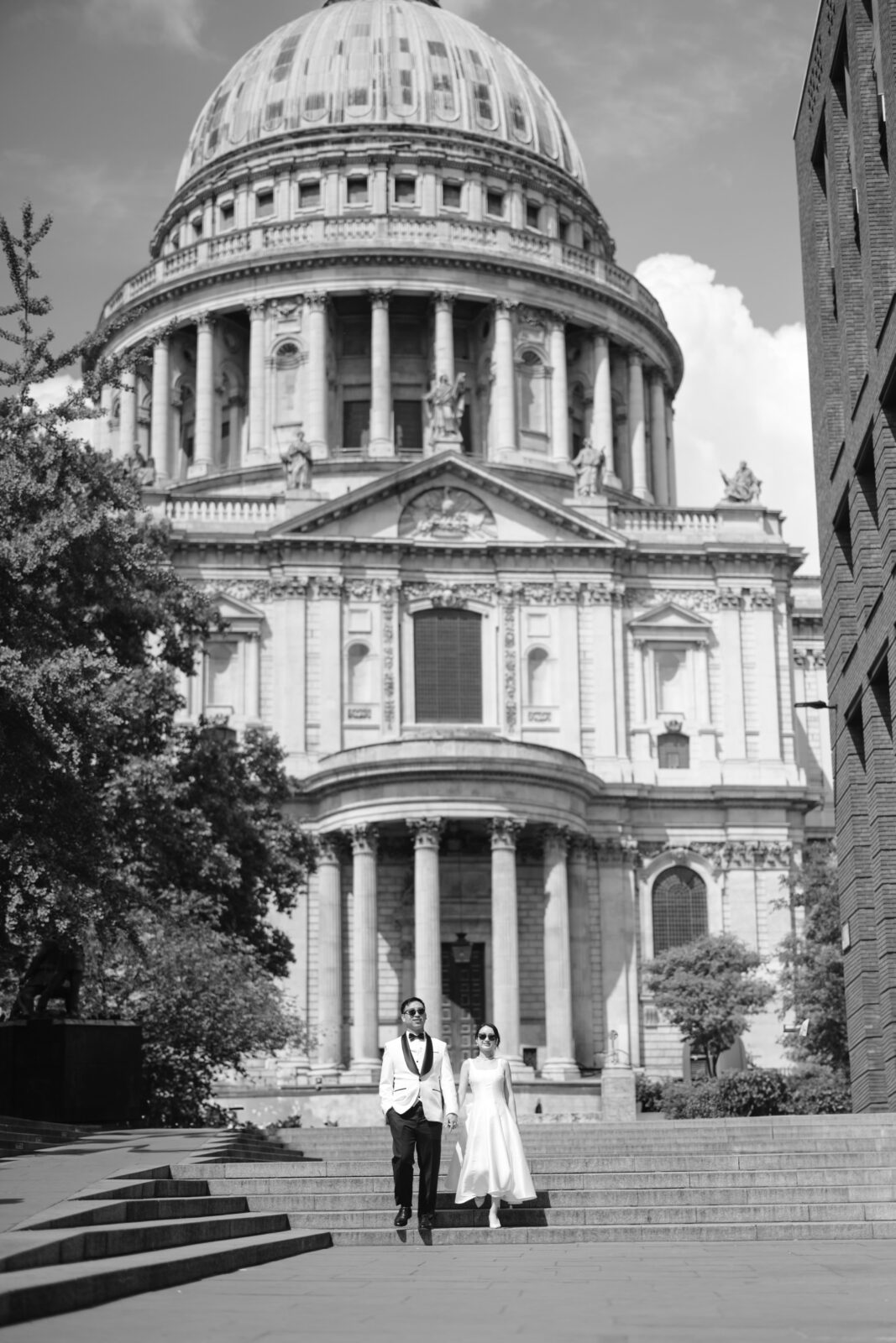 A couple dressed formally, with the man in a suit and the woman in a white dress, walk hand in hand outside St. Paul’s Cathedral in London on a sunny day.