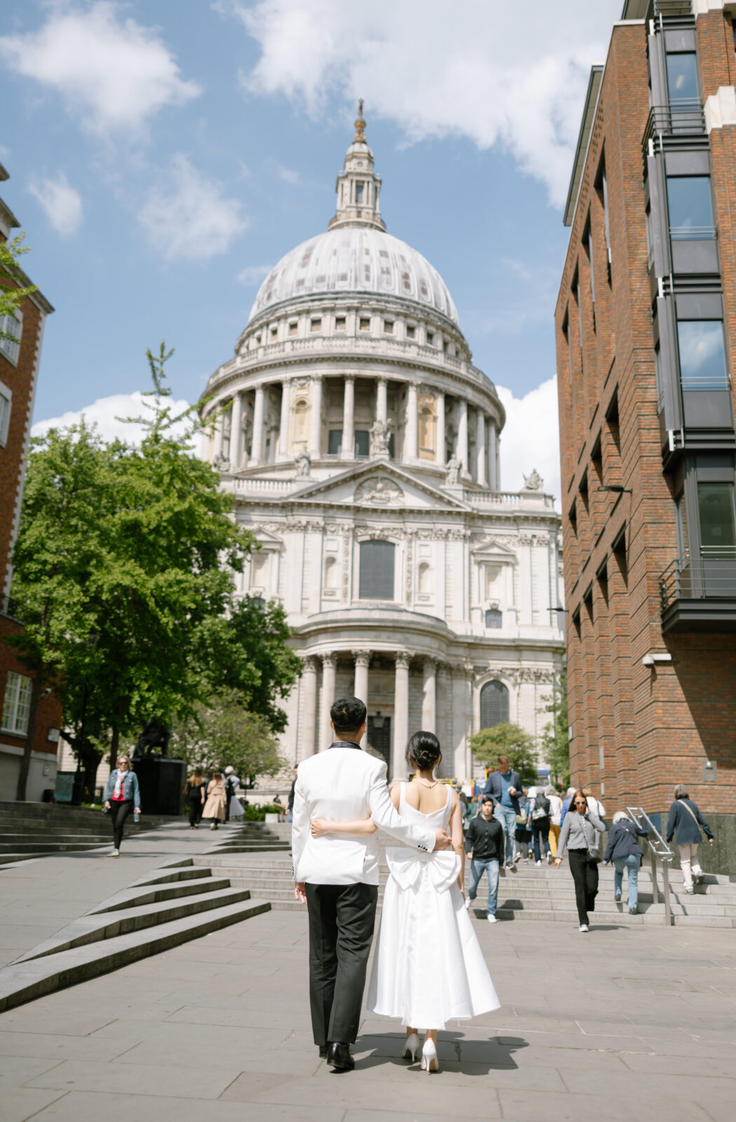 A couple in formal attire walks arm in arm toward St. Paul’s Cathedral in London on a sunny day, surrounded by people and framed by trees and brick buildings.