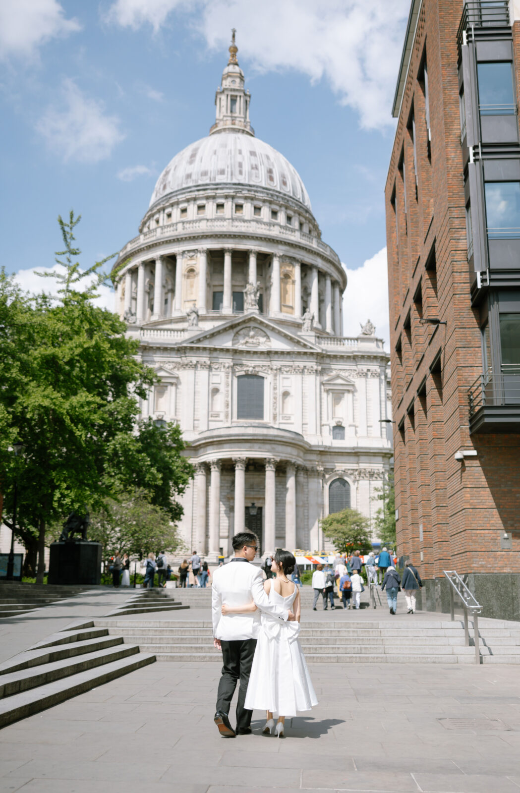 A couple dressed in white walks arm in arm toward St Pauls Cathedral in London on a sunny day, with crowds and trees in the background and modern brick buildings to the right.