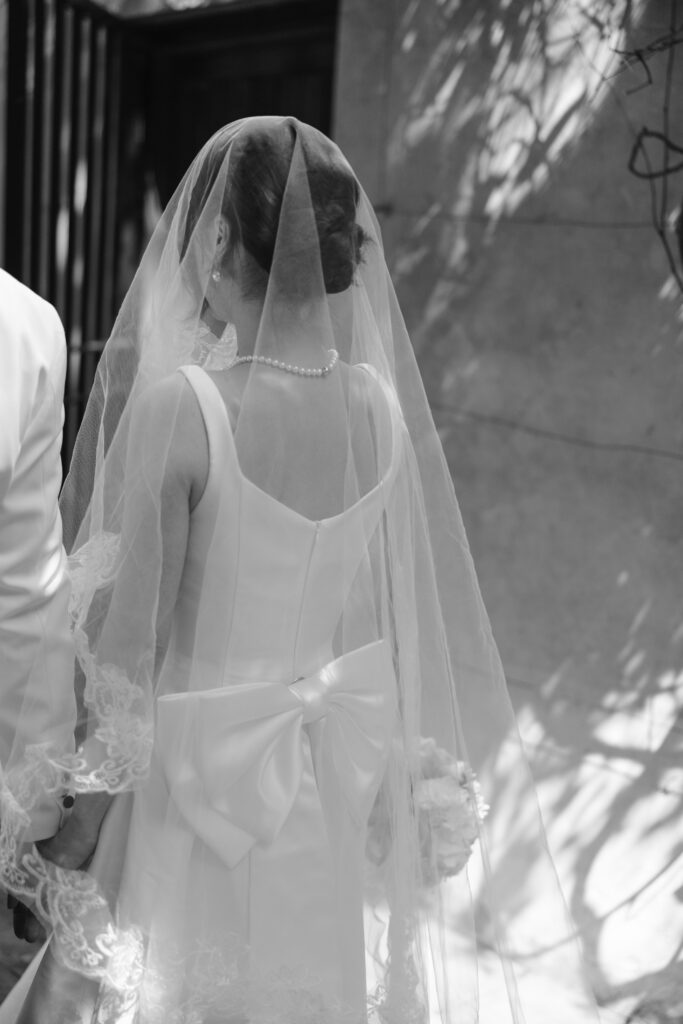 A bride in a white gown and veil, adorned with a pearl necklace and a large bow on the back of her dress, stands holding hands with someone, facing away, in a softly lit outdoor setting.