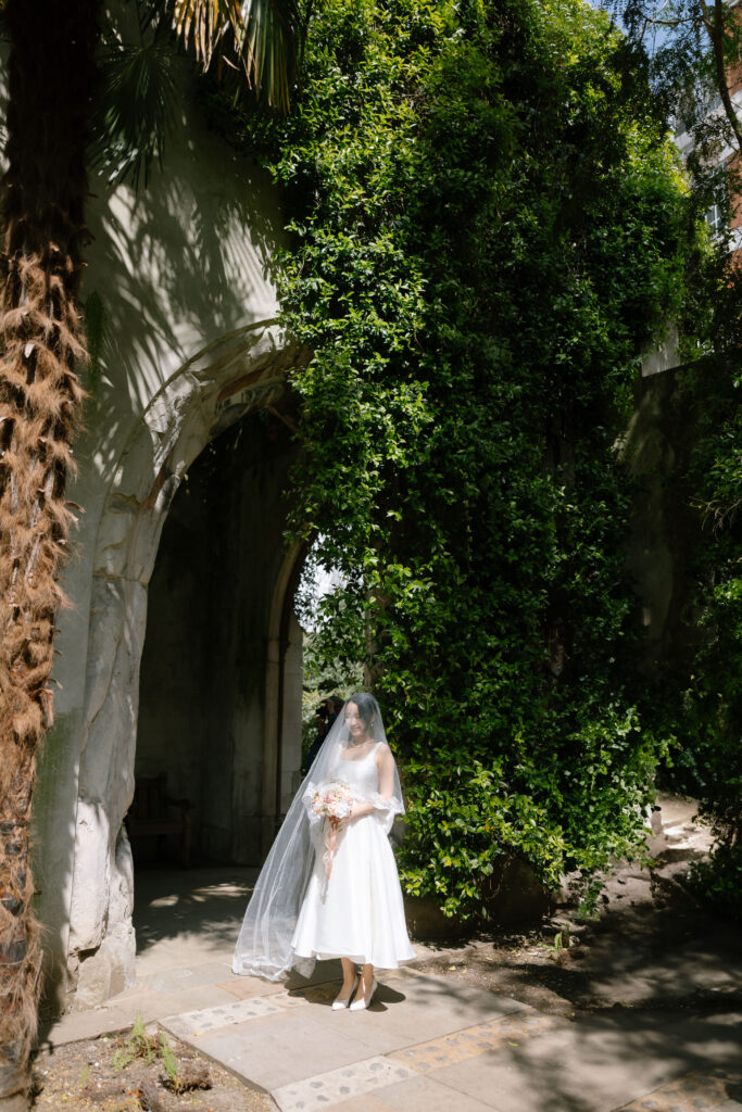A bride in a white dress and veil stands holding a bouquet under a stone archway covered with lush green ivy and bathed in sunlight.