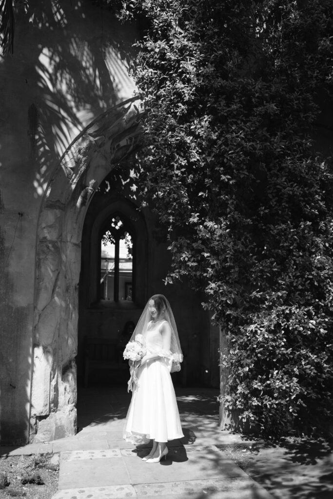 A bride in a white dress and veil stands in sunlight near a leafy archway outside an old stone building, holding a bouquet of flowers. The scene is in black and white.