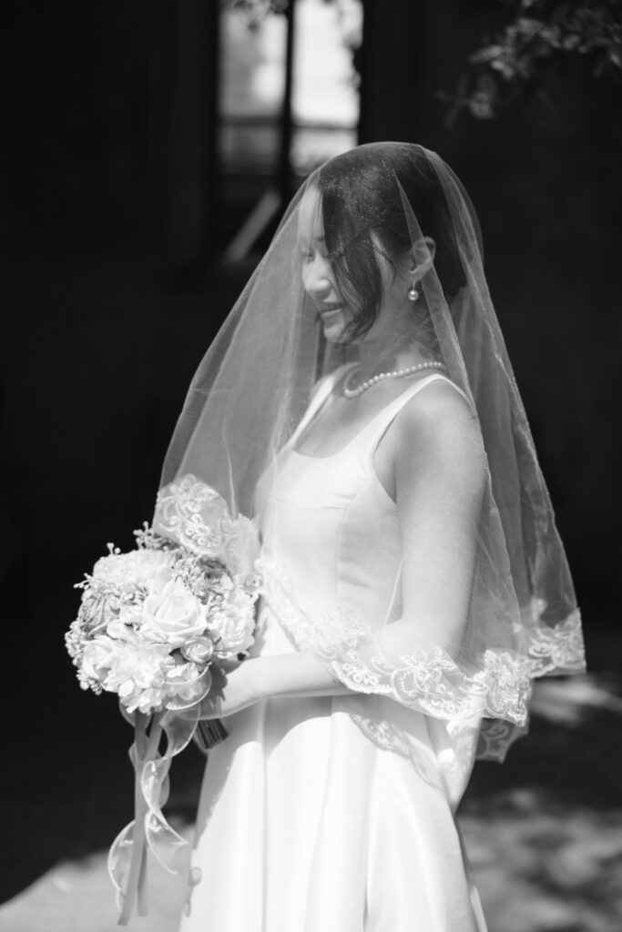 A bride in a white dress and veil stands holding a bouquet of flowers, smiling gently. The image is in black and white, with soft light highlighting her face and dress.