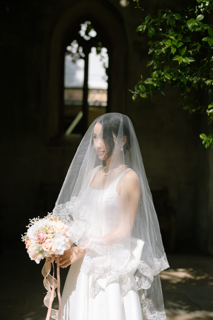 A bride in a white dress and lace veil stands indoors, holding a bouquet of flowers. Sunlight shines on her, with shadows and green leaves visible overhead. A tall arched window is in the background.
