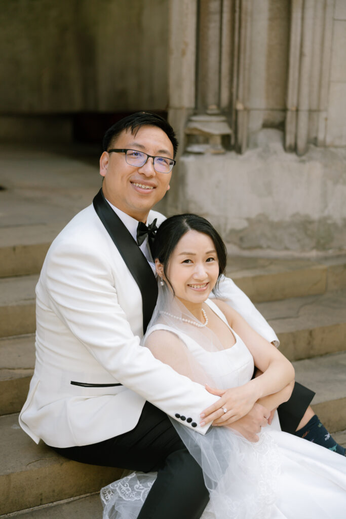 A smiling couple in wedding attire sits on stone steps. The groom wears a white tuxedo jacket and glasses, while the bride wears a white dress and veil. They embrace, looking happily at the camera.