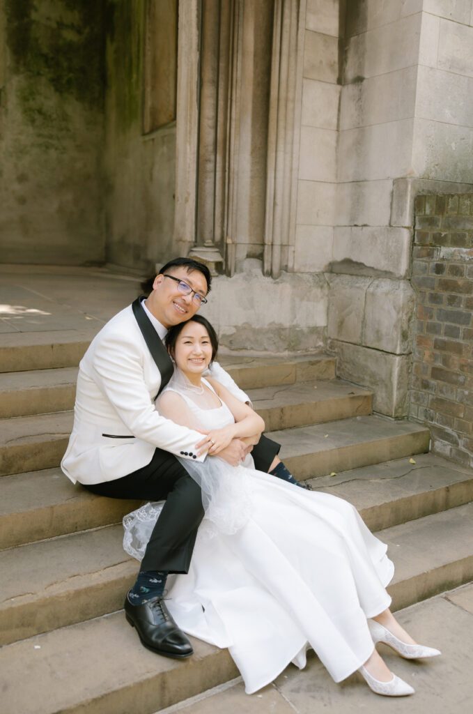 A smiling couple in wedding attire sits on stone steps, embracing. The groom wears a white tuxedo jacket and black pants, while the bride wears a white dress. They are seated by an old stone building with a relaxed, joyful expression.