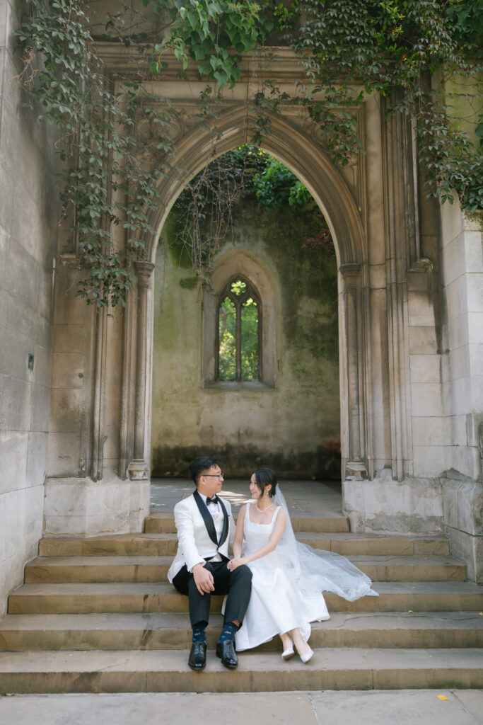 A newly married couple in formal wedding attire sits on stone steps beneath a gothic archway, surrounded by aged stone walls and green ivy, gazing at each other lovingly.