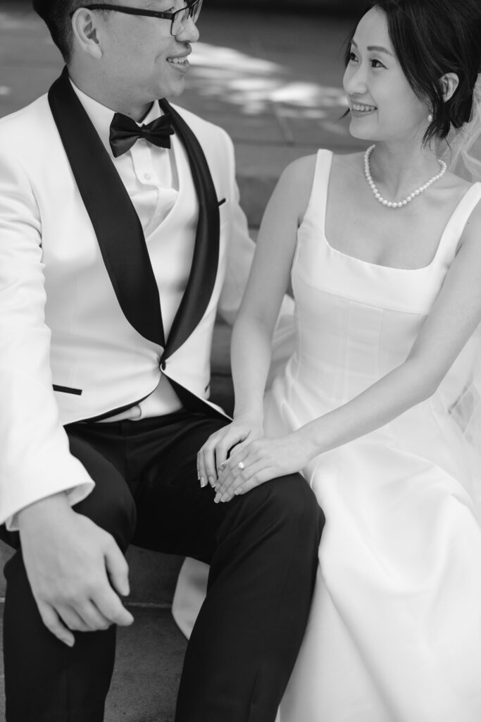 A bride and groom sit side by side, smiling at each other. The groom wears a white tuxedo jacket with a black lapel, and the bride wears a white dress and pearl necklace. They hold hands, looking happy.