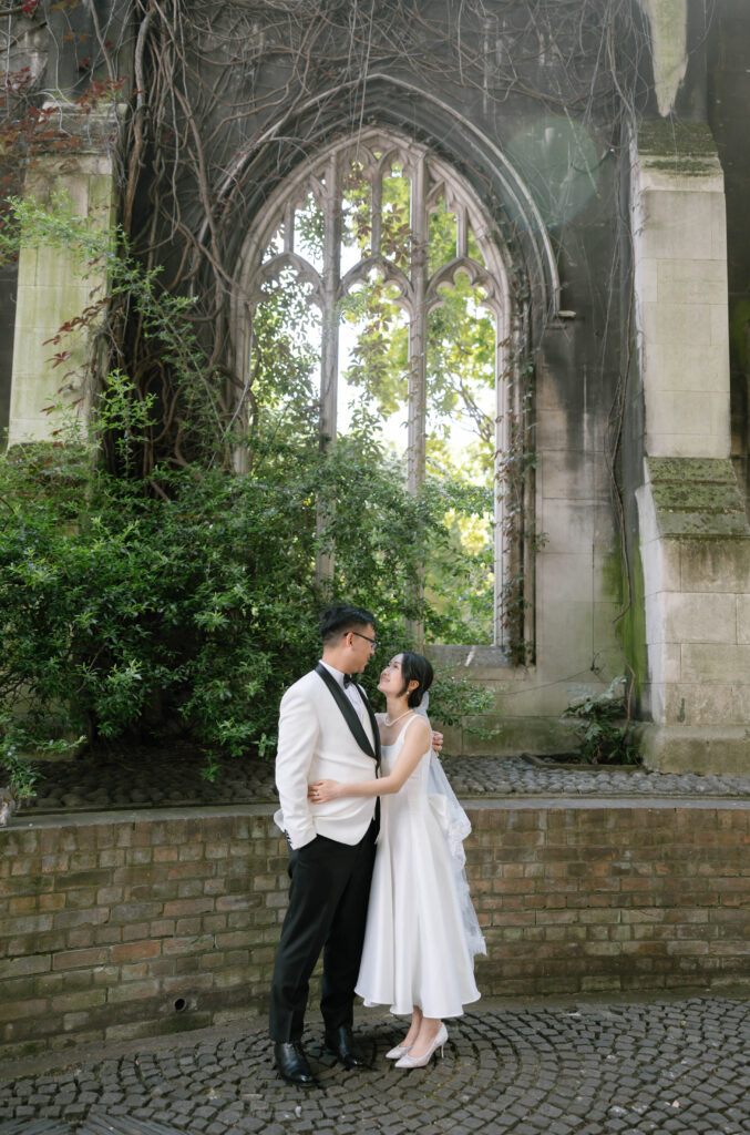 A couple in wedding attire stands closely together, gazing at each other, in front of a large, arched stone window overgrown with vines and surrounded by greenery.