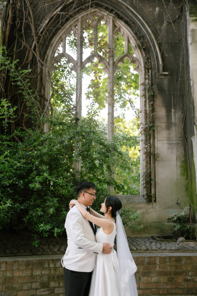 A bride and groom in formal attire embrace and look at each other in front of a large, arched stone window covered in greenery and vines, surrounded by old brick and lush plants.