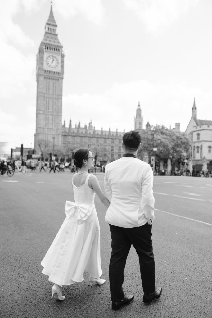 A couple dressed in formal white attire walks hand-in-hand across a street near Big Ben in London. The scene is in black and white, capturing a romantic and elegant moment with historic architecture in the background.