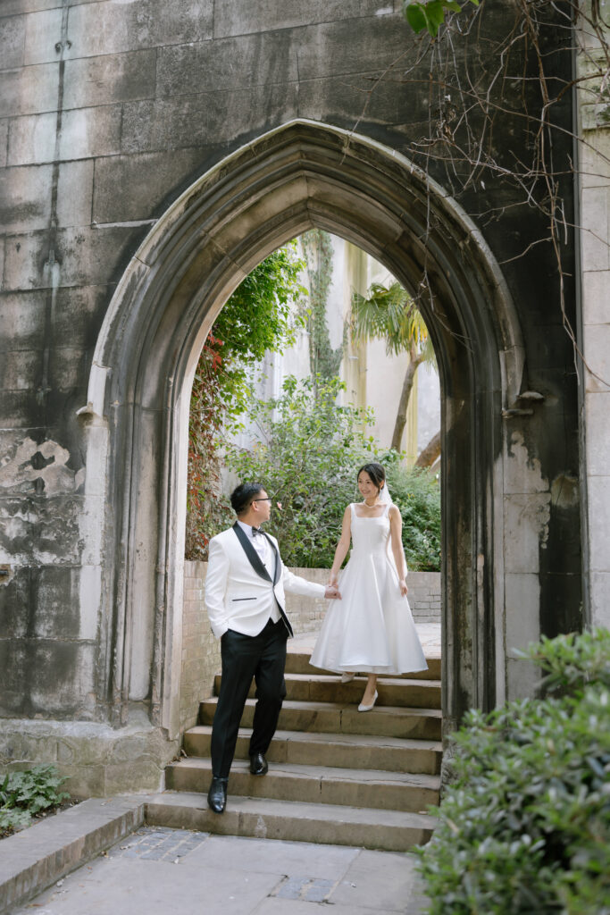 A bride and groom walk hand in hand under a stone archway, surrounded by greenery. The bride wears a white dress and the groom wears a white tuxedo jacket with black pants. They look at each other and smile.