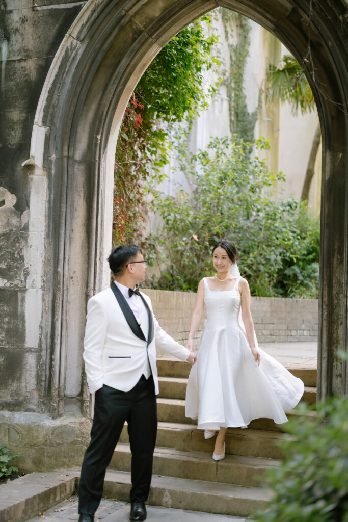 A couple dressed in formal attire, with the woman in a white dress and the man in a white jacket and black pants, hold hands while walking down stone steps under an old stone archway surrounded by greenery.