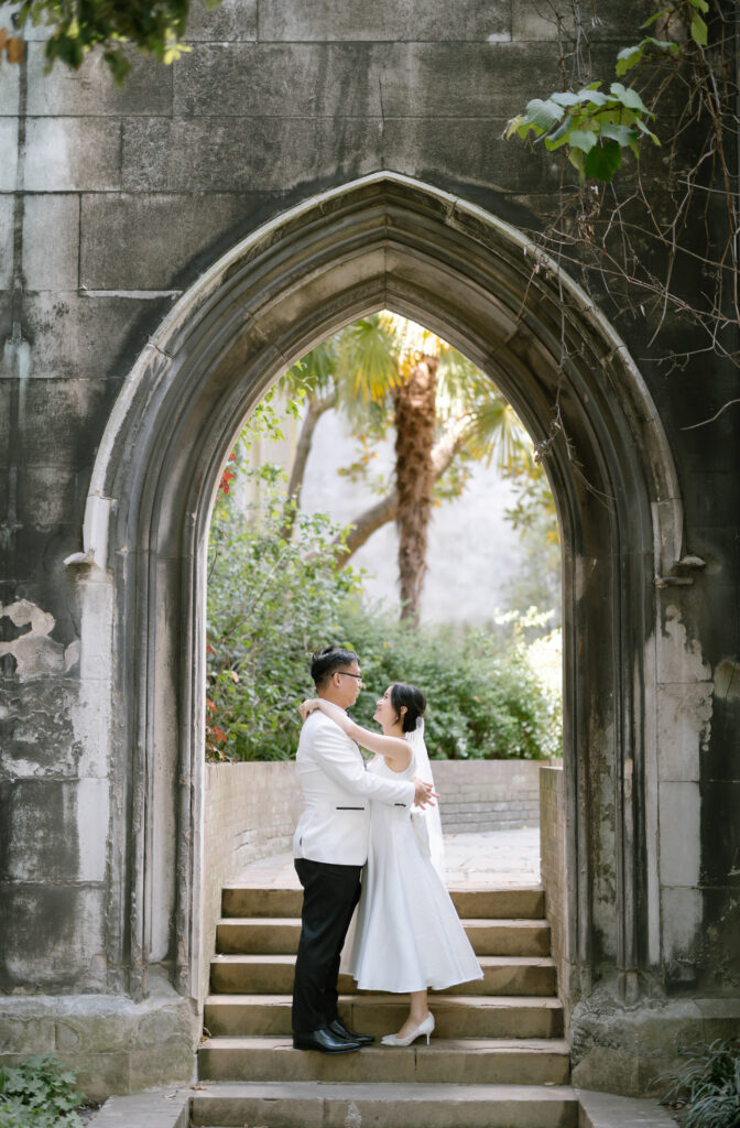 A bride and groom in wedding attire embrace on stone steps under a large, gothic archway in a garden setting, surrounded by greenery and sunlight.