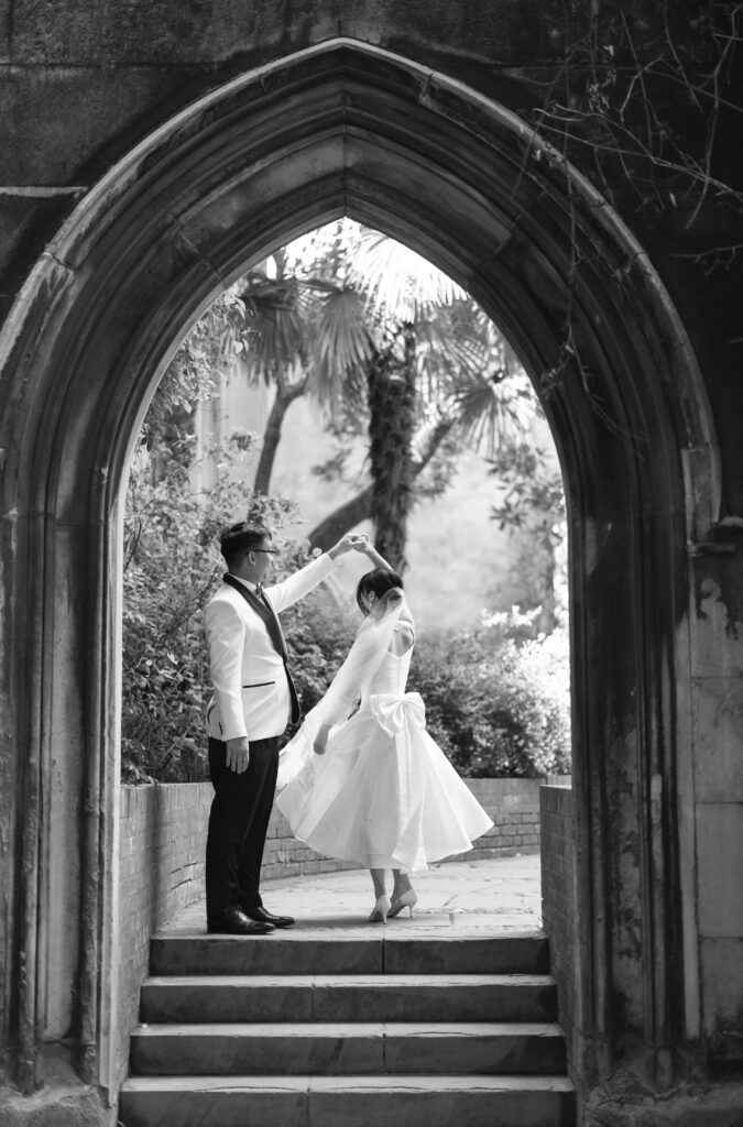 A couple dressed in formal attire dance under a stone archway surrounded by lush greenery. The woman twirls in a white dress and the man, in a light jacket and dark pants, holds her hand. The scene is captured in black and white.