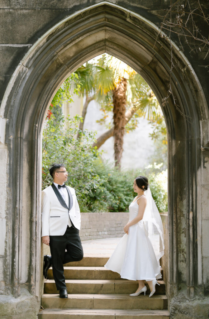 A bride in a white dress and veil stands facing a groom in a white tuxedo jacket and black pants under a stone archway, surrounded by greenery and sunlight.