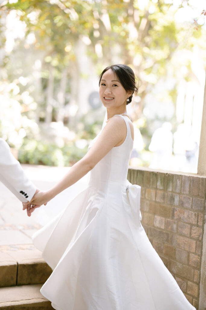A woman in a white wedding dress smiles while holding someones hand and turning back, outdoors with sunlight filtering through trees in the background.