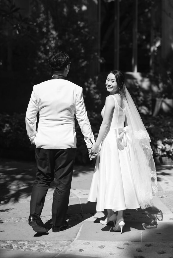 A bride in a white dress and veil smiles at the camera while holding hands with a groom in a white jacket. They are walking outdoors on a sunny day.