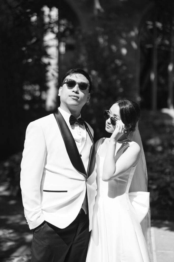 A bride and groom in formal wedding attire, both wearing sunglasses, pose confidently outdoors in bright sunlight. The groom wears a white tuxedo with a black lapel; the bride wears a sleeveless gown and veil.
