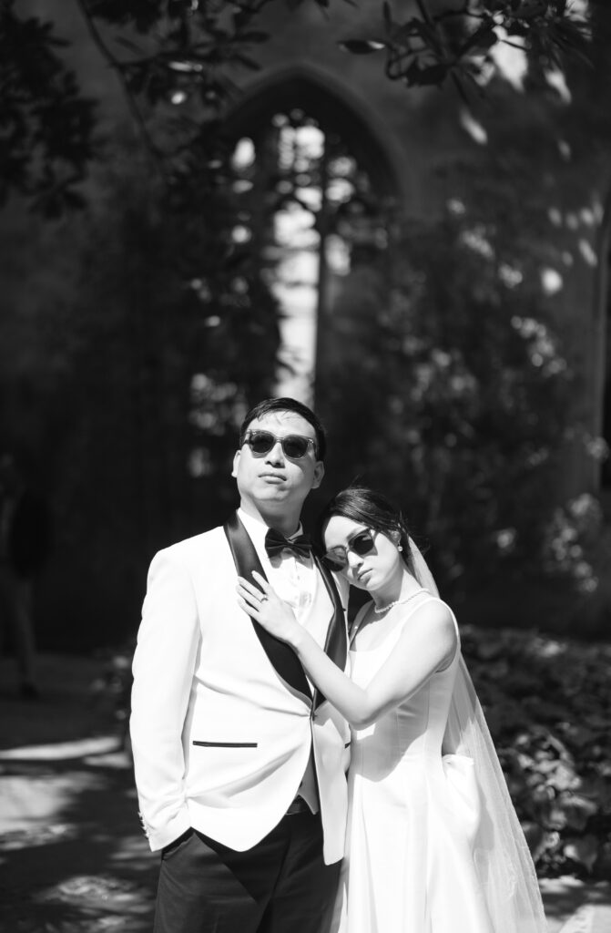 A bride and groom wearing sunglasses pose outdoors. The groom wears a white tuxedo with a black bow tie, and the bride in a sleeveless gown rests her head on his shoulder, both standing under dappled sunlight.