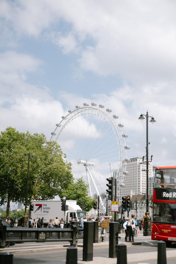 A view of the London Eye Ferris wheel on a partly cloudy day, with trees, a double-decker red bus, and city buildings in the foreground. Pedestrians and vehicles are visible along the street.