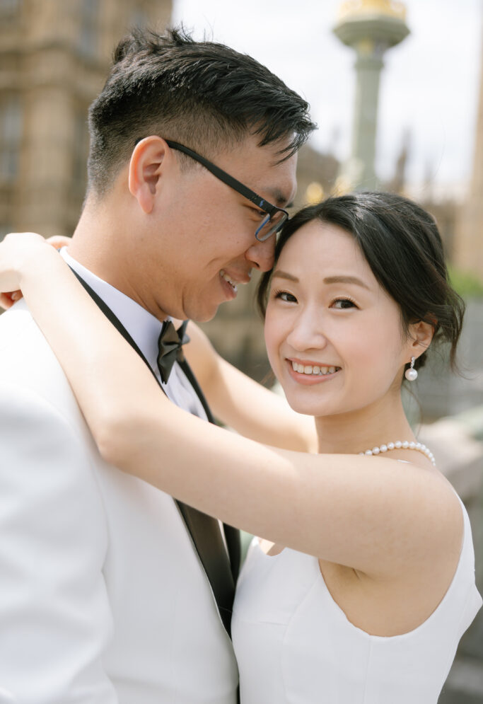 A couple dressed in formal attire embrace and smile outdoors. The woman wears a white dress with a pearl necklace; the man wears glasses, a white jacket, and a black bow tie. A historic building is blurred in the background.