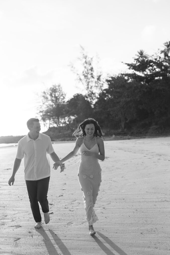 A couple walks barefoot on a sandy beach, holding hands and smiling. The woman wears a long dress and the man wears a short-sleeved shirt and pants. Trees and soft sunlight are visible in the background.
