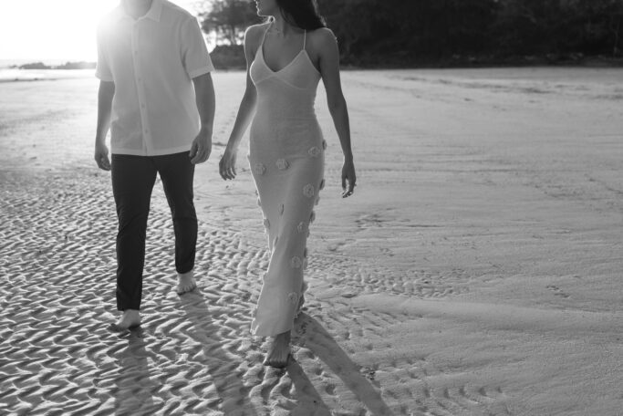 A man and woman walk barefoot on a sandy beach at sunset, wearing light, summery clothes. The image is in black and white, and they appear relaxed, enjoying the peaceful shoreline together.