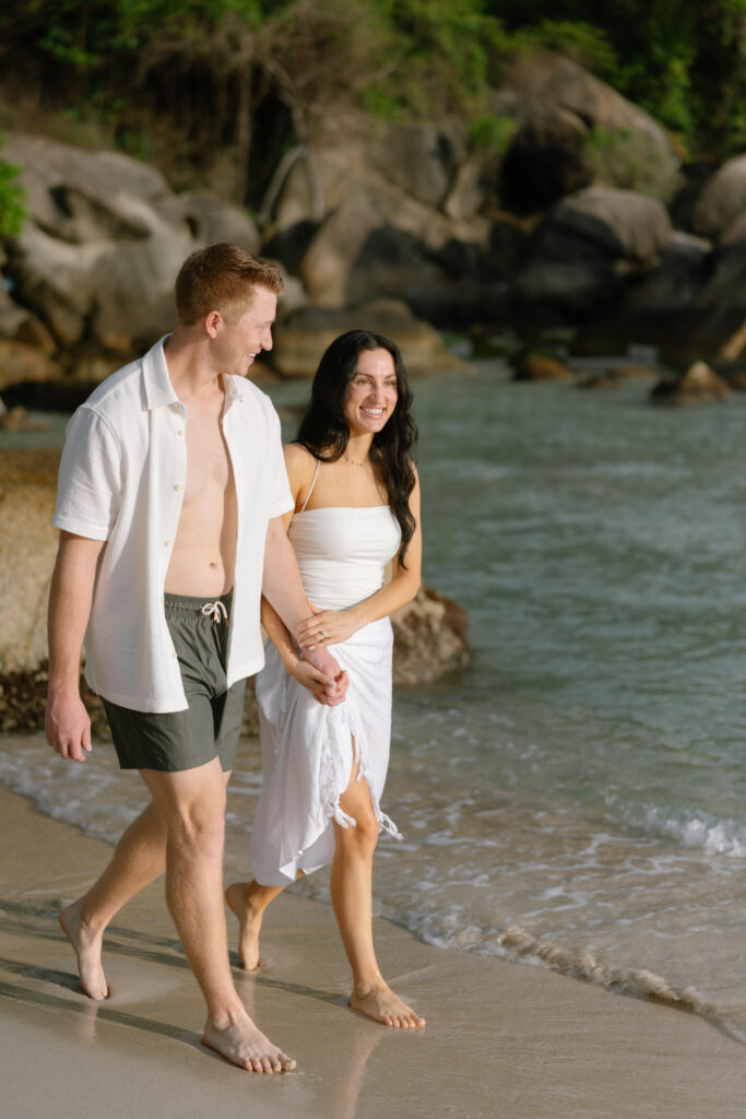 A couple walks barefoot along the shoreline, holding hands and smiling. The man wears swim shorts and an open shirt; the woman wears a white dress. The beach is sandy with rocks and greenery in the background.