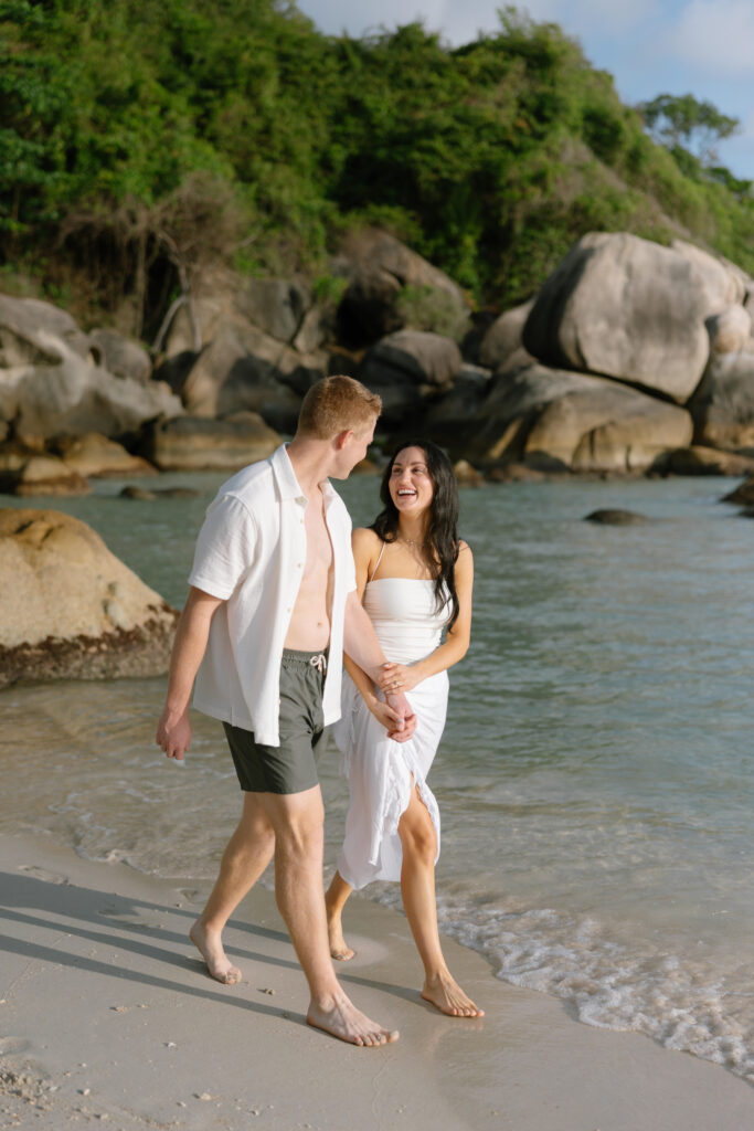 A smiling couple walks barefoot along a sandy beach near large rocks and greenery, holding hands and looking at each other under the sunlight.