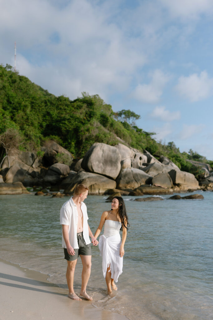 A couple walks barefoot along a sandy beach, holding hands and smiling at each other. Behind them are large rocks, green foliage, and a partly cloudy sky.