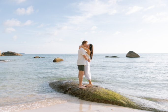 A couple embraces and kisses on a mossy rock at the edge of the sea, with gentle waves, sandy shore, and blue sky in the background.