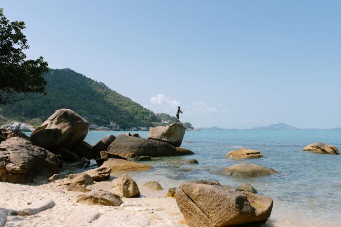 A person stands on a large rock by the sea, surrounded by scattered boulders and clear, shallow water. In the background, there are green hills and distant islands under a blue sky.