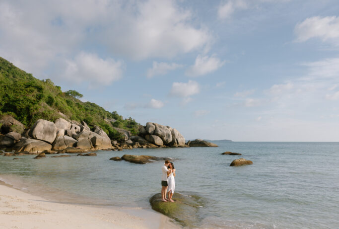 A couple embraces on a rock by the shoreline, surrounded by clear water, sandy beach, large boulders, and lush green hills under a partly cloudy blue sky.