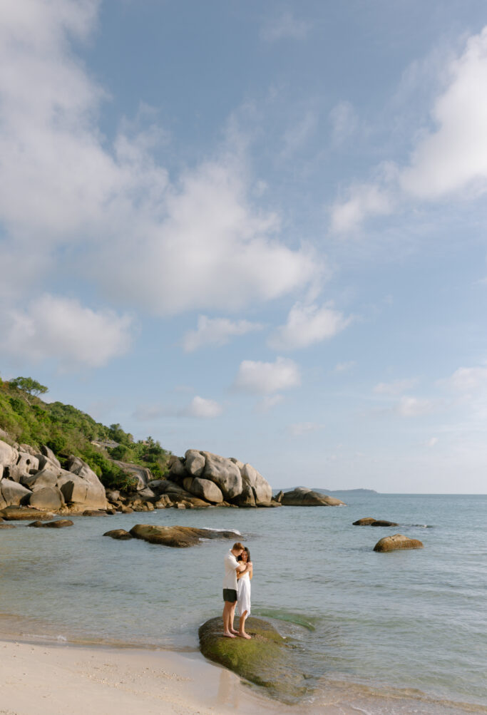 A couple stands on a large rock at the edge of the sea, embracing under a bright sky with scattered clouds. Rocky shoreline and greenery are visible in the background.