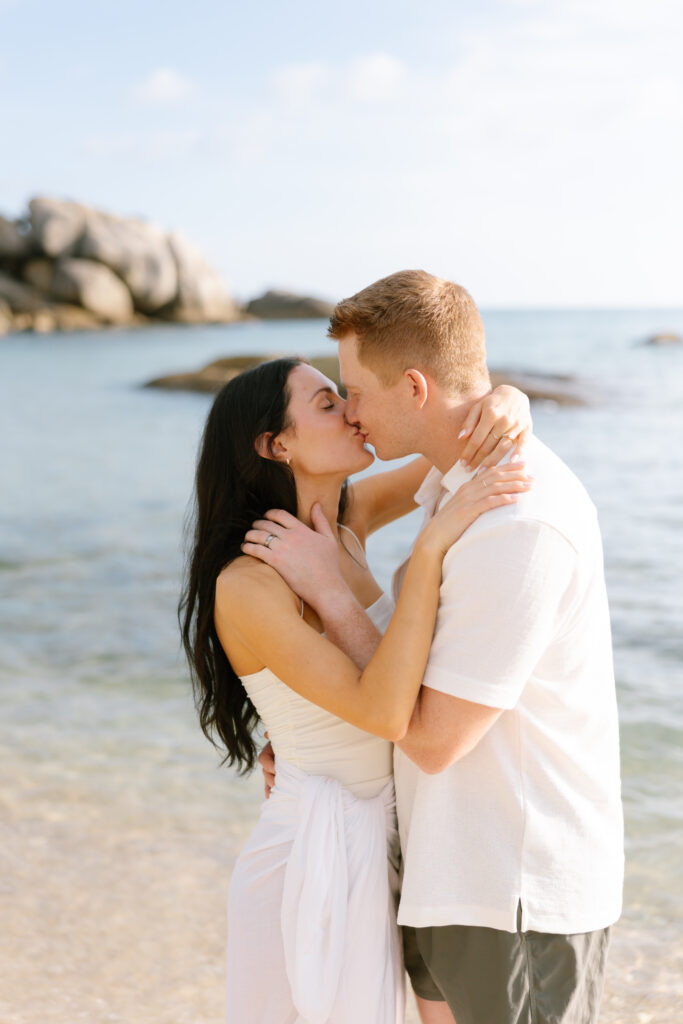 A couple stands on a sandy beach, embracing and kissing near the shoreline. The woman has long dark hair and wears a white dress, while the man has short hair and wears a light shirt. Rocks and calm water are in the background.