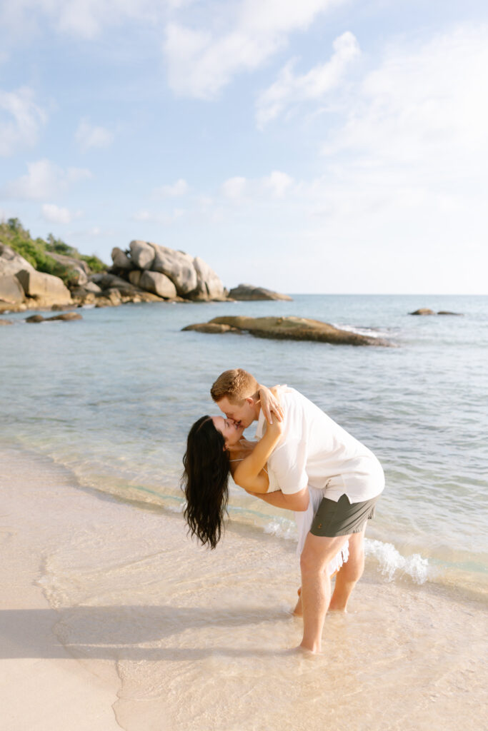 A couple stands in shallow water on a sandy beach, with the man dipping the woman as they kiss. Rocky formations and blue sky are in the background. Both appear happy and relaxed, enjoying the sunny day.